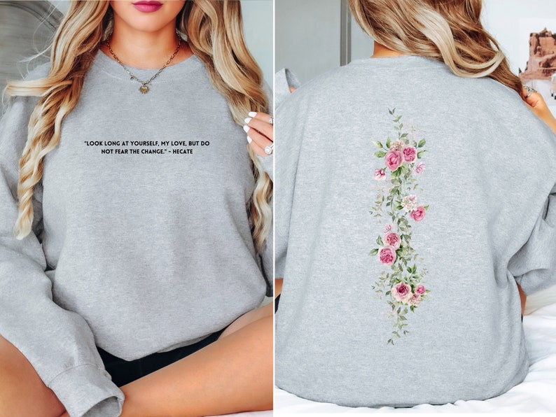 K&ouml;nnte beinhalten: Grauer Pullover mit einem Zitat auf der Vorderseite und einem Blumendesign auf der R&uuml;ckseite. Das Zitat lautet "Schau lange auf dich selbst, meine Liebe, aber f&uuml;rchte dich nicht vor dem Wandel - Hekate".