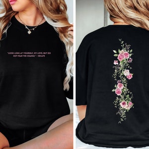 K&ouml;nnte beinhalten: Schwarzes Oversize-T-Shirt mit einem rosafarbenen Blumendesign auf der R&uuml;ckseite und einem Zitat in rosafarbener Schrift auf der Vorderseite: "LOOK LONG AT YOURSELF, MY LOVE, BUT DO NOT FEAR THE CHANGE. - HECATE"