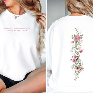 K&ouml;nnte beinhalten: Wei&szlig;er Pullover mit einem rosafarbenen Zitat auf der Vorderseite, das lautet "Schau lange auf dich selbst, meine Liebe, aber f&uuml;rchte dich nicht vor der Ver&auml;nderung." - Meg Cabot. Die R&uuml;ckseite des Pullovers zeigt ein vertikales Blumenmuster mit rosa Rosen und gr&uuml;nen Bl&auml;ttern.