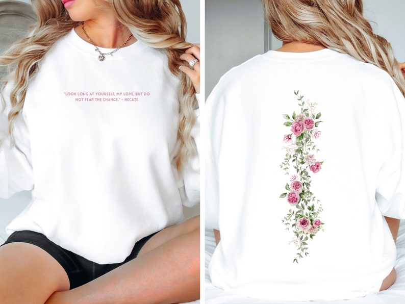 K&ouml;nnte beinhalten: Wei&szlig;er Pullover mit einem rosafarbenen Zitat auf der Vorderseite, das lautet "Schau lange auf dich selbst, meine Liebe, aber f&uuml;rchte dich nicht vor der Ver&auml;nderung." - Meg Cabot. Die R&uuml;ckseite des Pullovers zeigt ein vertikales Blumenmuster mit rosa Rosen und gr&uuml;nen Bl&auml;ttern.