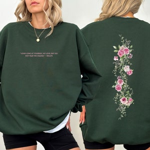 K&ouml;nnte beinhalten: Ein dunkelgr&uuml;ner Pullover mit einem rosafarbenen Blumendesign auf der R&uuml;ckseite und einem Zitat in rosafarbener Schrift auf der Vorderseite, das lautet: "Schau lange auf dich selbst, meine Liebe, aber f&uuml;rchte dich nicht vor der Ver&auml;nderung - Hekate"