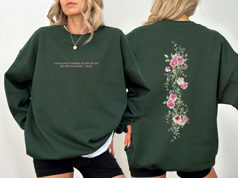 K&ouml;nnte beinhalten: Ein dunkelgr&uuml;ner Pullover mit einem rosafarbenen Blumendesign auf der R&uuml;ckseite und einem Zitat in rosafarbener Schrift auf der Vorderseite, das lautet: "Schau lange auf dich selbst, meine Liebe, aber f&uuml;rchte dich nicht vor der Ver&auml;nderung - Hekate"
