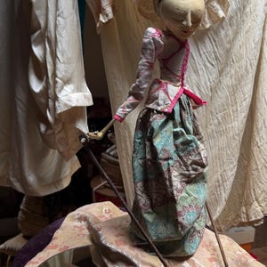 Peut inclure: Une marionnette à tige Wayang Golek avec une tête en bois sculpté au visage peint, vêtue d'une veste blanche à motifs à manches longues avec des bordures roses et une jupe volumineuse à motif batik floral vert, marron et bleu, tenue par deux tiges.