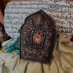 May include: An ornate, antique metal religious artifact. The dark metal frame surrounds a central image of a seated figure with a red robe. The frame is intricately detailed with relief carvings. The artifact rests on a draped, light-colored fabric.