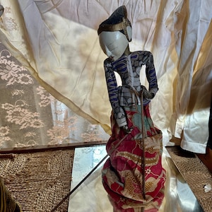 Op de afbeelding: Een traditionele Indonesische Wayang Golek-pop. De pop heeft een wit gezicht, een donkerblauwe en gouden hoofdtooi en is gekleed in een rood en blauw kostuum met patronen. Hij staat op een spiegeloppervlak.