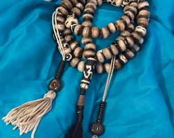 May include: A prayer bead necklace featuring round, weathered-look beads in shades of brown and cream, interspersed with carved skull-shaped beads. The necklace includes three dangling ends with dark tassels and a central bead marked with an Om symbol.