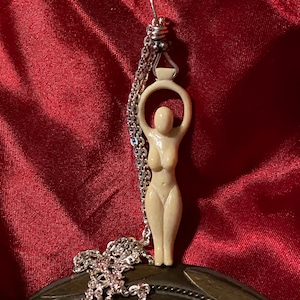 Può includere: Una collana con un ciondolo in ceramica bianca che rappresenta una figura femminile stilizzata con le braccia alzate, che tiene un cerchio sopra la sua testa. La collana è realizzata con una catena d'argento.