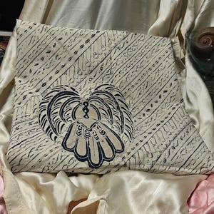 May include: Cream-colored fabric with a dark blue, stylized floral design. The fabric has a diagonal striped pattern. The design is centered on the fabric. The fabric is folded and resting on a satin-like surface.