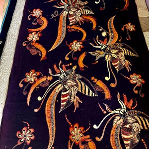 Op de afbeelding: Donker marineblauwe stof met een herhaald patroon van gestileerde bloemen en insectachtige ontwerpen in oranje, bruin en crème. Het patroon bevat lange, gebogen elementen en gedetailleerde bloemaccenten, waardoor een ingewikkeld textielontwerp ontstaat.