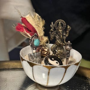 Peut inclure: Un bol blanc à bordure dorée rempli de sable et contenant divers petits objets spirituels. On y trouve une petite statue de divinité en laiton, une pierre yin-yang et une bague turquoise.