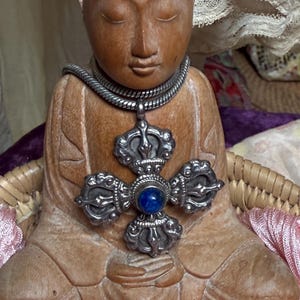 Op de afbeelding: Een houten Boeddha-standbeeld met een zilveren ketting en hanger. De hanger heeft een blauwe steen in het midden van een decoratief zilveren kruis. Het beeld zit in een meditatieve houding.