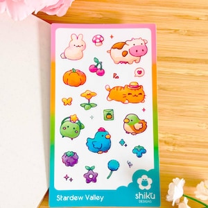 Puede incluir: Una hoja de pegatinas coloridas con ilustraciones de animales, plantas y alimentos. Las pegatinas incluyen un conejo, una vaca, un gato, una calabaza, cerezas y más. El texto "Stardew Valley" y "shiku DESIGNS" está impreso en la parte inferior.