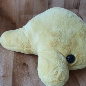 May include: A plush yellow manatee toy with black eyes. The toy is lying on a wooden surface.