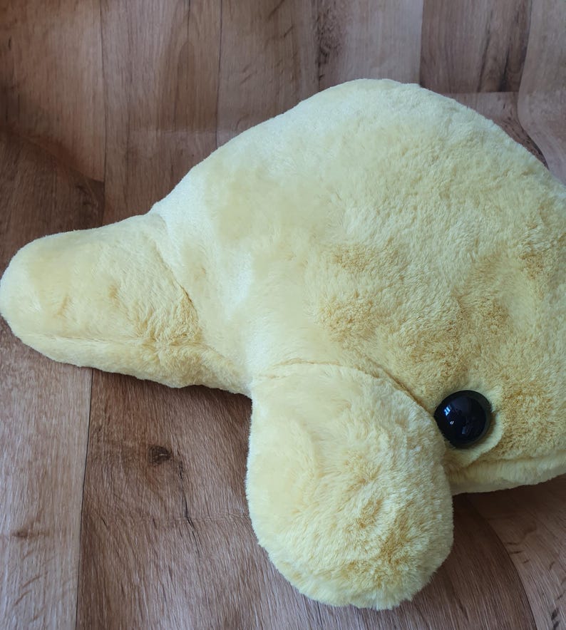 May include: A plush yellow manatee toy with black eyes. The toy is lying on a wooden surface.