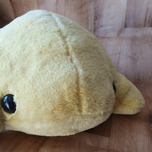 May include: A plush yellow stuffed toy with black eyes. The toy has a seam down the centre of its back.