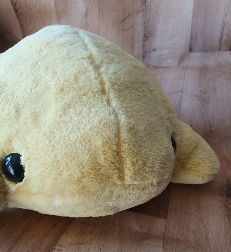 May include: A plush yellow stuffed toy with black eyes. The toy has a seam down the centre of its back.