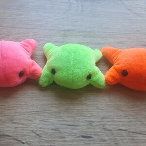 May include: Three plush toys shaped like frogs in pink, green, and orange colors. The toys have black eyes and are lying on a wooden surface.