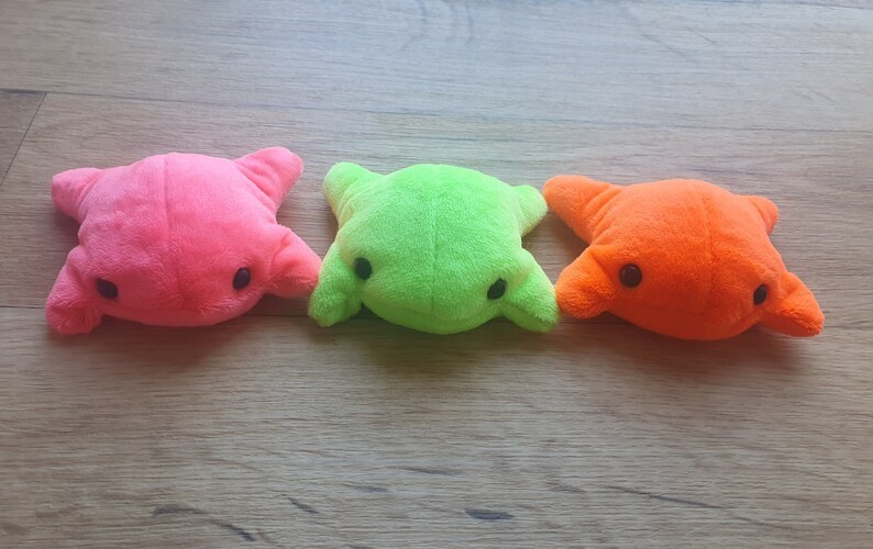 May include: Three plush toys shaped like frogs in pink, green, and orange colors. The toys have black eyes and are lying on a wooden surface.