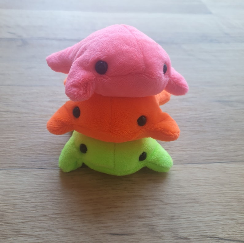 May include: A stack of three plush toys shaped like dolphins. The top dolphin is pink, the middle dolphin is orange, and the bottom dolphin is green.