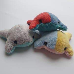 May include: Three plush toys shaped like stingrays in different colors. The first stingray is gray with a light blue underside. The second stingray is red, blue, and yellow. The third stingray is blue, yellow, and pink.