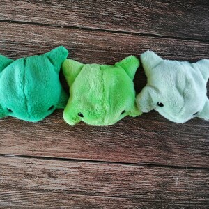Pu&ograve; includere: Tre giocattoli di peluche a forma di creatura dei cartoni animati con un gradiente di colore verde, verde chiaro e grigio. I giocattoli sono disposti in fila su una superficie di legno.