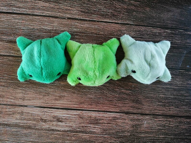 Pu&ograve; includere: Tre giocattoli di peluche a forma di creatura dei cartoni animati con un gradiente di colore verde, verde chiaro e grigio. I giocattoli sono disposti in fila su una superficie di legno.