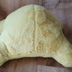 May include: A soft, yellow plush turtle-shaped cushion. The cushion is made of a fluffy, fuzzy material and has a slightly textured surface.