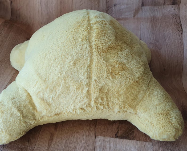 May include: A soft, yellow plush turtle-shaped cushion. The cushion is made of a fluffy, fuzzy material and has a slightly textured surface.