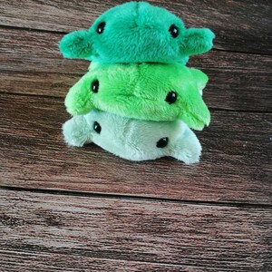 Pu&ograve; includere: Tre giocattoli di peluche impilati uno sopra l'altro. Il giocattolo in cima &egrave; di colore verde brillante, il giocattolo nel mezzo &egrave; di colore verde chiaro e il giocattolo in fondo &egrave; di colore verde pallido. Tutti e tre i giocattoli hanno occhi neri e una forma semplice e arrotondata.