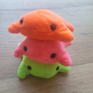 May include: Three plush toys stacked on top of each other. The top toy is orange, the middle toy is pink, and the bottom toy is green. Each toy has two black eyes.