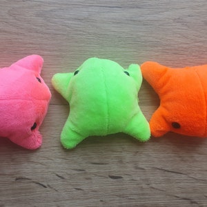 May include: Three plush toys shaped like stars in pink, green, and orange. The toys are lying on a wooden surface.