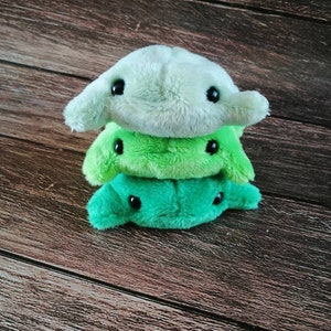 Pu&ograve; includere: Tre giocattoli di peluche impilati uno sopra l'altro. Il giocattolo superiore &egrave; verde chiaro, il giocattolo intermedio &egrave; verde scuro e il giocattolo inferiore &egrave; verde brillante. Tutti e tre i giocattoli hanno occhi neri e una forma semplice e arrotondata.