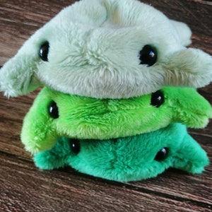 Pu&ograve; includere: Tre giocattoli di peluche impilati uno sopra l'altro. Il giocattolo superiore &egrave; verde chiaro, il giocattolo intermedio &egrave; verde brillante e il giocattolo inferiore &egrave; verde scuro. Tutti e tre i giocattoli hanno occhi neri e una forma semplice e arrotondata.
