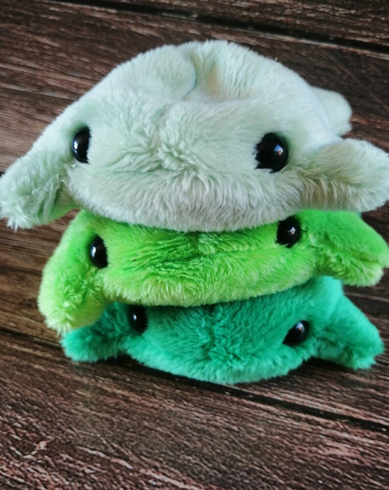 Pu&ograve; includere: Tre giocattoli di peluche impilati uno sopra l'altro. Il giocattolo superiore &egrave; verde chiaro, il giocattolo intermedio &egrave; verde brillante e il giocattolo inferiore &egrave; verde scuro. Tutti e tre i giocattoli hanno occhi neri e una forma semplice e arrotondata.