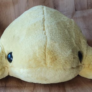 May include: A plush yellow dolphin toy with black eyes. The dolphin is lying on its belly with its flippers outstretched.