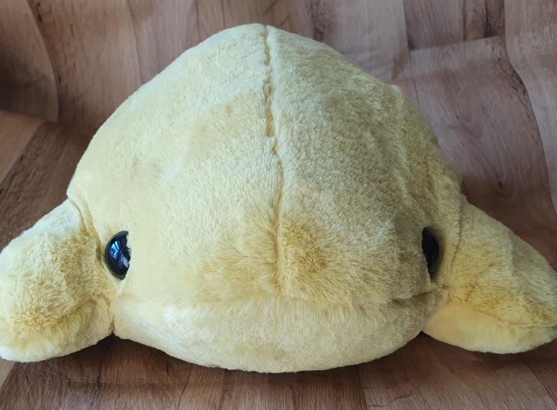 May include: A plush yellow dolphin toy with black eyes. The dolphin is lying on its belly with its flippers outstretched.