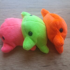 May include: Three plush toys in the shape of dolphins. The dolphins are pink, green, and orange. They are all facing the same direction and are lined up in a row.