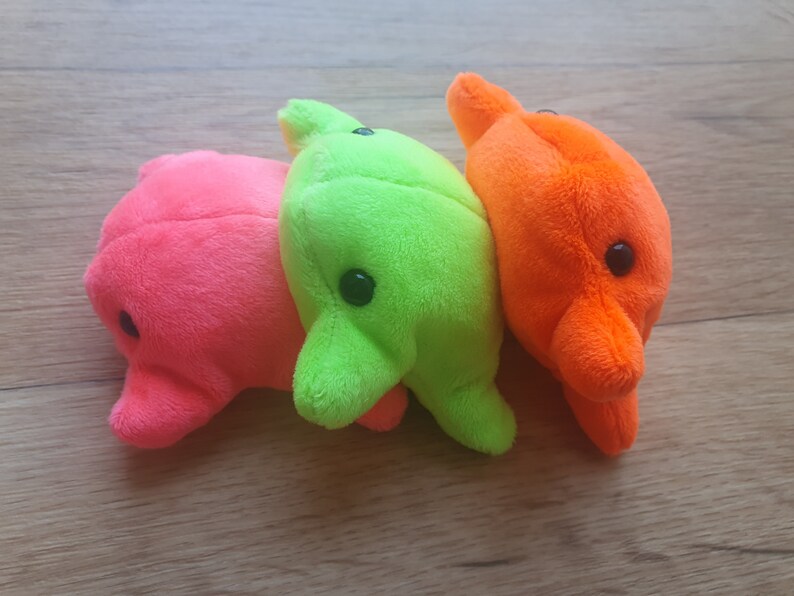May include: Three plush toys in the shape of dolphins. The dolphins are pink, green, and orange. They are all facing the same direction and are lined up in a row.
