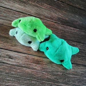 Pu&ograve; includere: Tre giocattoli di peluche a forma di razza in tonalit&agrave; di verde. I giocattoli hanno occhi neri e sono disposti in una pila su una superficie di legno.