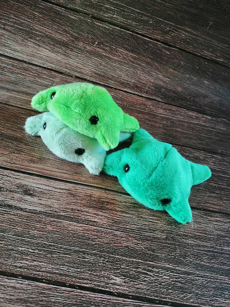 Pu&ograve; includere: Tre giocattoli di peluche a forma di razza in tonalit&agrave; di verde. I giocattoli hanno occhi neri e sono disposti in una pila su una superficie di legno.