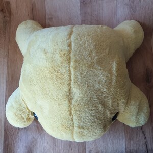 May include: A large, yellow plush toy shaped like a turtle. The toy has a soft, fuzzy texture and is lying on a wooden surface.
