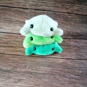 Pu&ograve; includere: Tre giocattoli di peluche impilati uno sopra l'altro. Il giocattolo in cima &egrave; bianco con occhi neri. Il giocattolo nel mezzo &egrave; verde chiaro con occhi neri. Il giocattolo in basso &egrave; verde scuro con occhi neri.