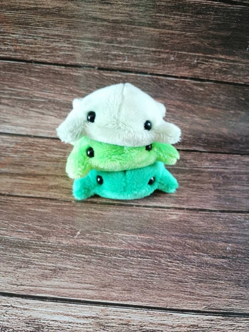 Pu&ograve; includere: Tre giocattoli di peluche impilati uno sopra l'altro. Il giocattolo in cima &egrave; bianco con occhi neri. Il giocattolo nel mezzo &egrave; verde chiaro con occhi neri. Il giocattolo in basso &egrave; verde scuro con occhi neri.