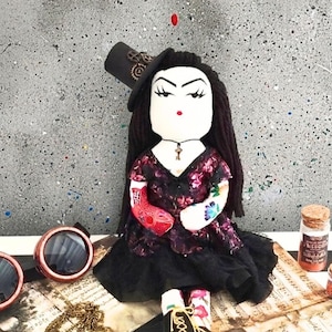 Könnte beinhalten: Handgefertigte Puppe mit schwarzen Haaren, die einen schwarzen Zylinder mit Zahnrädern, ein Blumenkleid und einen Schlüsselanhänger trägt. Die Puppe hat Tätowierungen an den Armen und sitzt auf Notenblättern.