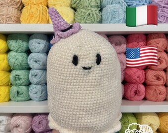 May include: White crocheted ghost amigurumi wearing a purple witch hat. The ghost has black eyes and a smiling mouth. The ghost is sitting on a hand in front of a shelf of colorful yarn.