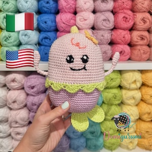 May include: A pink crocheted amigurumi toy with a yellow flower on its head. The toy has a smiling face with black eyes and a pink mouth. The toy is holding up its arms and has green crocheted feet. The toy is in front of a background of colorful yarn balls.