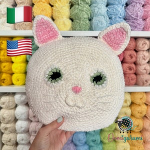 May include: White crocheted cat head pillow with pink ears, green eyes, and a pink nose. The pillow is surrounded by colorful yarn balls.