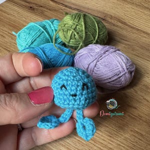 Può includere: Un amigurumi di medusa all'uncinetto blu fatto a mano con un volto sorridente e tentacoli pendenti. La medusa è tenuta in mano, con diversi gomitoli di filato nei toni del blu, verde e viola sullo sfondo. L'immagine include il testo "Domigurumi".