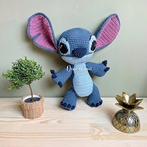 Può includere: Bambola Stitch blu all'uncinetto fatta a mano con grandi orecchie, interno rosa e occhi neri. La bambola è in posa con le braccia tese. Sono visibili anche una piccola pianta in vaso e un oggetto dorato a forma di ananas.