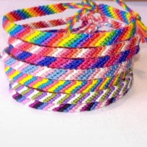 Könnte beinhalten: Ein Stapel bunter Freundschaftsarmbänder. Die Armbänder bestehen aus gewebten Fäden in verschiedenen Farben, darunter Rot, Orange, Gelb, Grün, Blau und Lila. Die Armbänder sind etwa 1,3 cm breit.